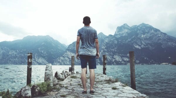 man standing on gray concrete dock facing body of water and mountains at daytime